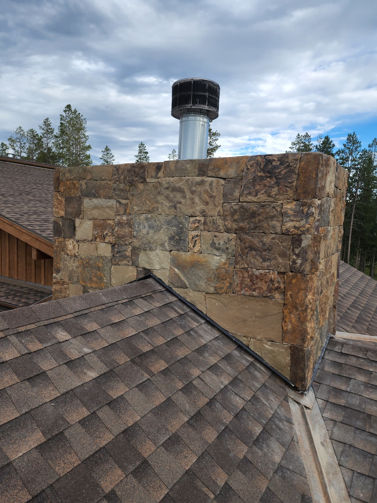 Squared-stone chimney chase at a shingle roofline against pines and sky